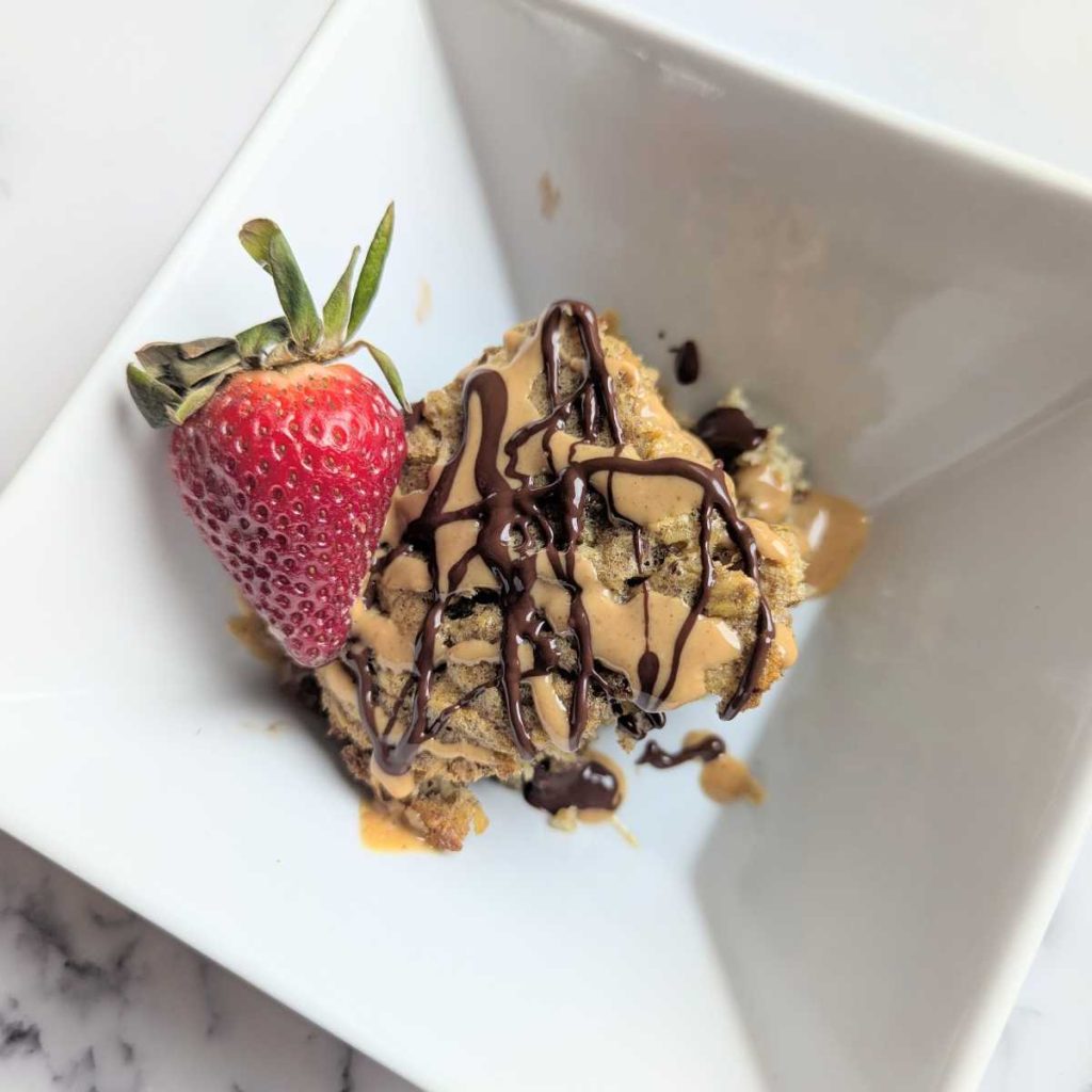 baked protein oats in bowl with peanut butter and chocolate drizzle with a strawberry on the side