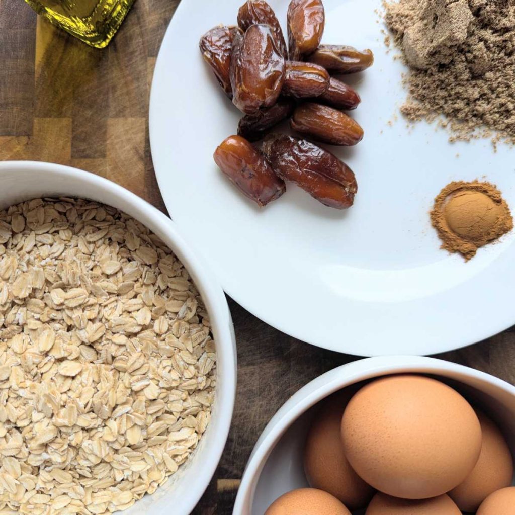 separate ingredients laid out on a cutting board including oatmeal, banana, eggs, olive oil, cinnamon, dates, flax seed meal
