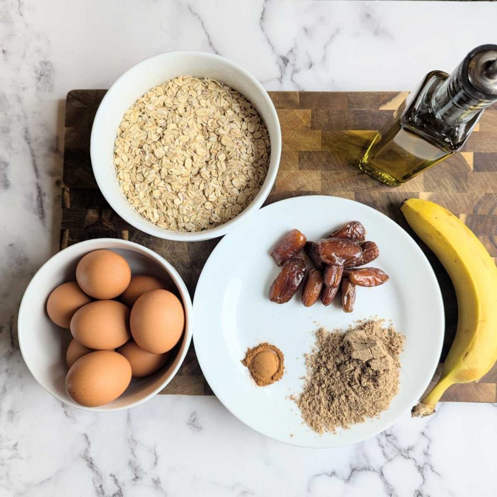 separate ingredients laid out on a cutting board including oatmeal, banana, eggs, olive oil, cinnamon, dates, flax seed meal